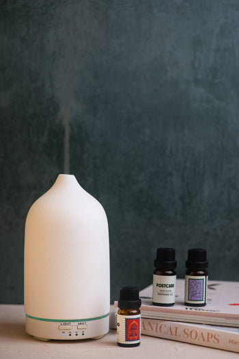 A white Postcard Moon Bloom diffuser oil (10g) sits beside three small bottles and two stacked books, set against a dark green background—evoking a serene lunar lounge ambiance.