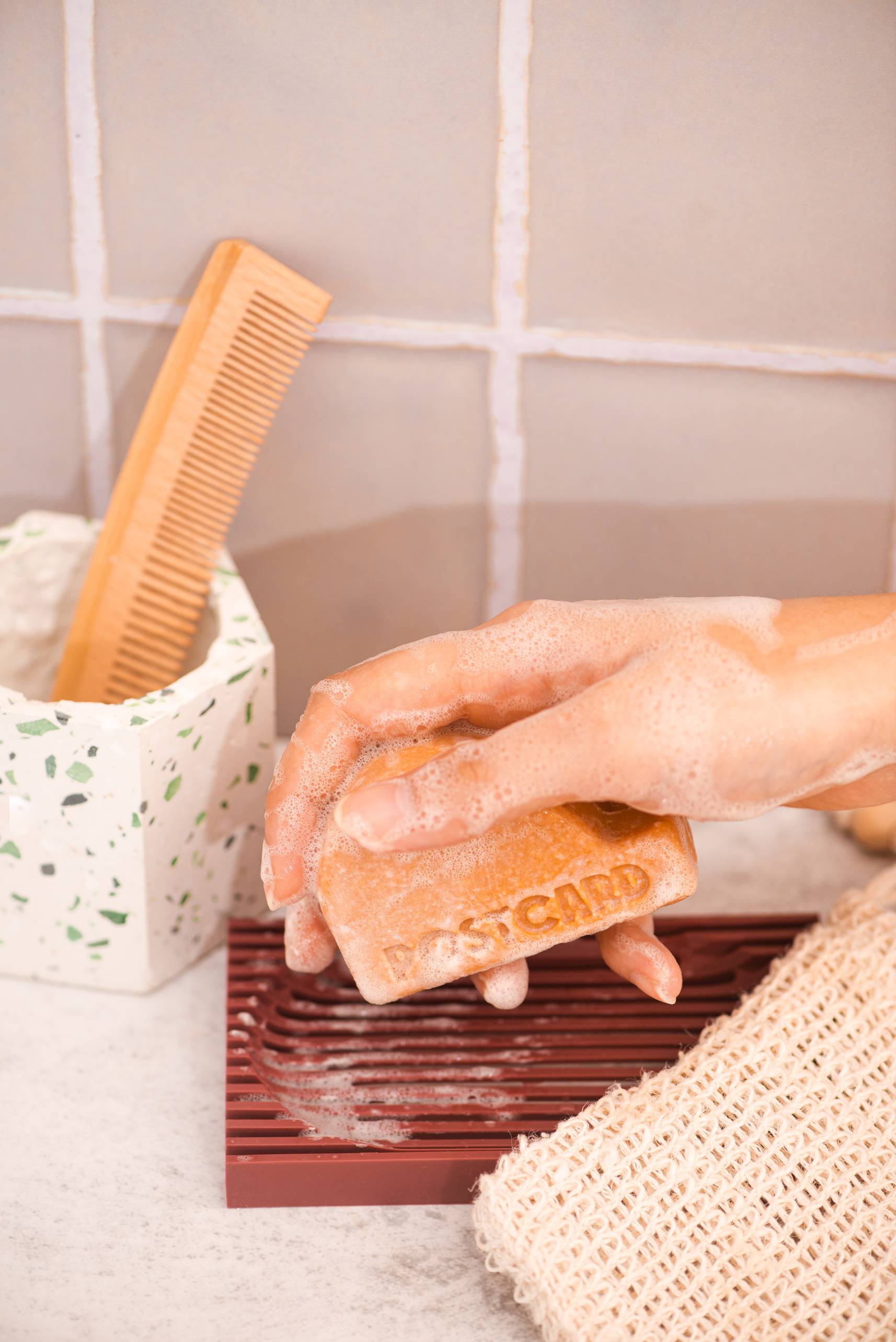A hand covered in suds holds the Postcard Head-To-Toe Bar, 100g - Citrus Circus over a soap dish, with a wooden comb and textured white jar beside a woven washcloth. This sulfate-free bar by Postcard energizes your routine.