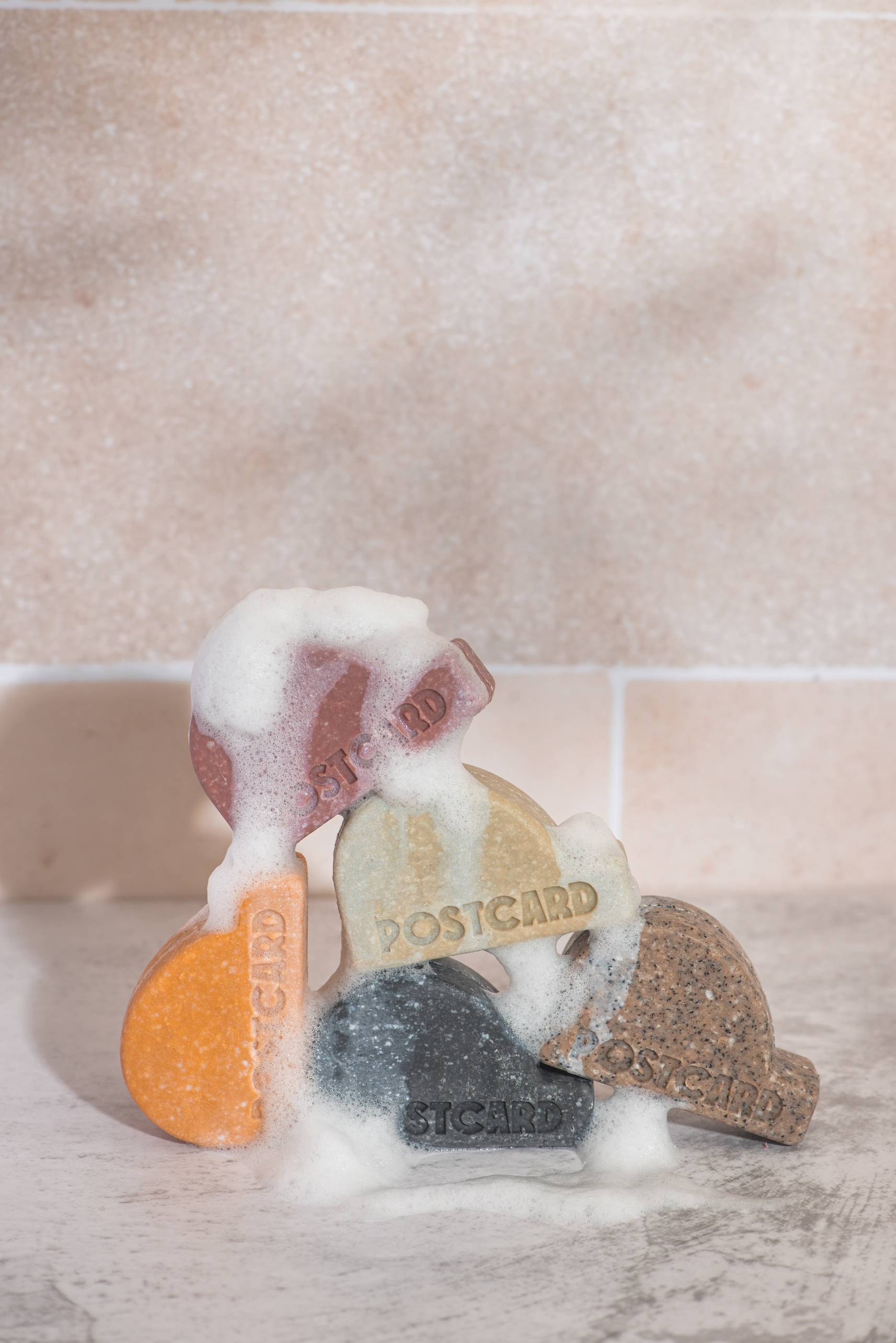 Five colorful, rounded Postcard Head-To-Toe Bars (Icy Peaks, 100g) are stacked on a light pink tiled surface, with white suds covering parts of these sulfate-free, all-in-one bars from Postcard.