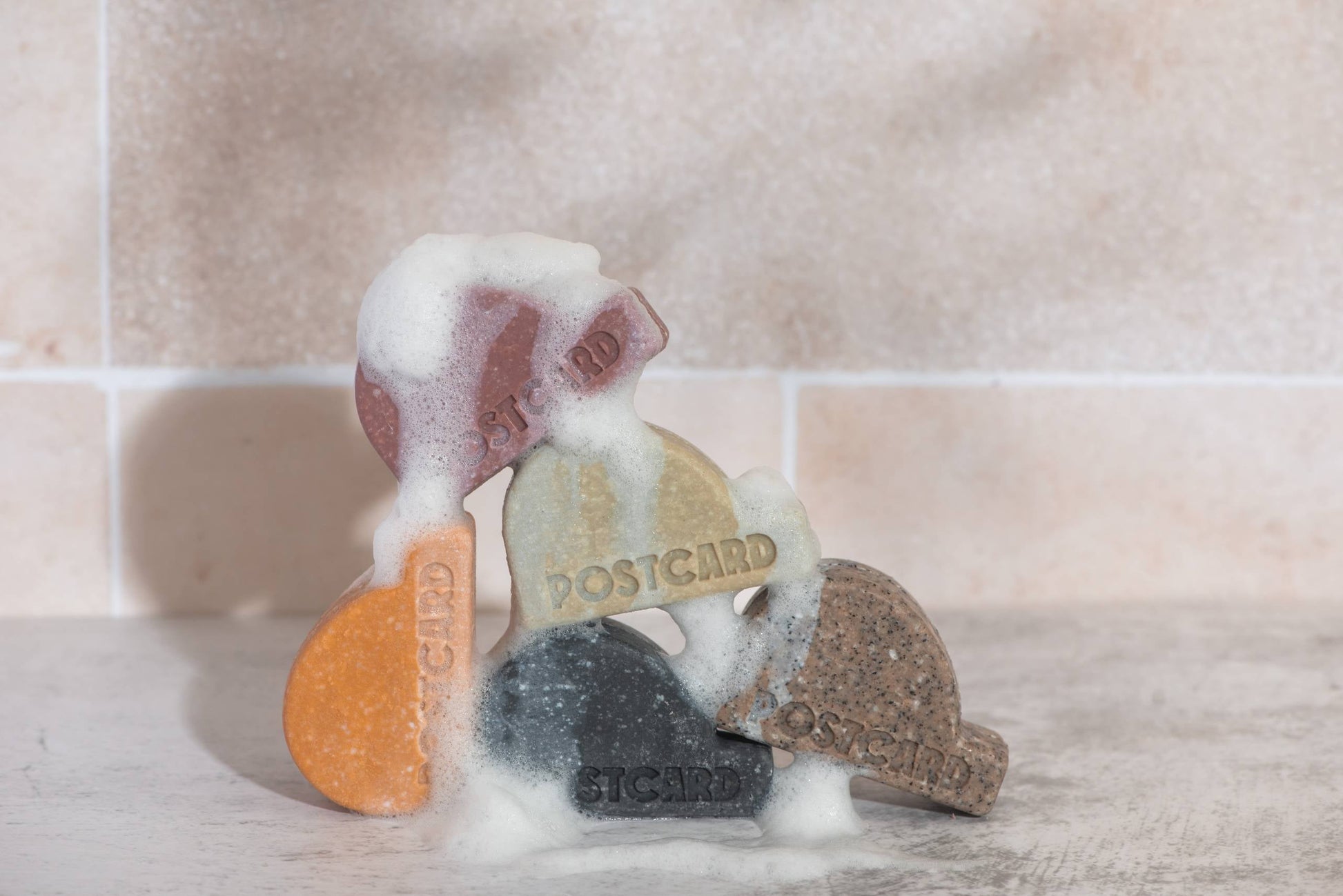 Five colorful, half-moon POSTCARD Head-To-Toe Bars (100g - Icy Peaks) are stacked on a tiled surface with white suds, set against a light-tiled wall. Each sulfate-free bar from Postcard is suitable for all-in-one cleansing.