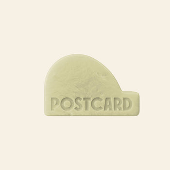 A pale yellow, rounded rectangular Postcard Head-To-Toe Bar (100g, Java Jungle) with POSTCARD embossed in uppercase on a beige background—an all-in-one, sulfate-free bar designed to combat hair loss.