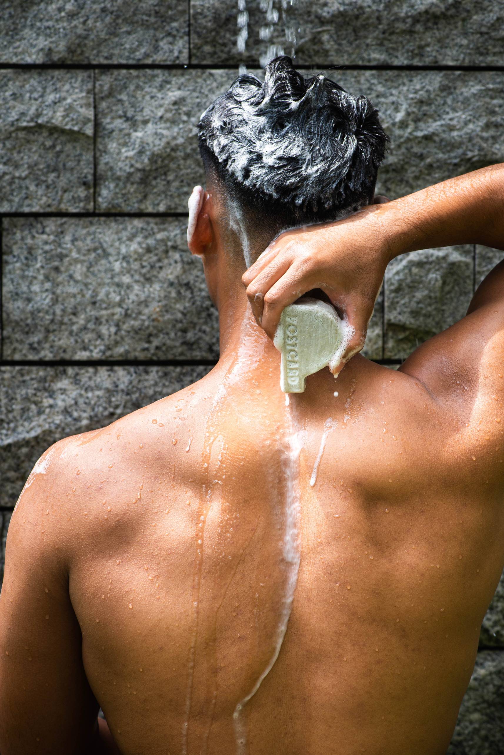 A person with short dark hair is showering, seen from behind, using the Postcard Head-To-Toe Bar, 100g - Java Jungle by Postcard to wash their upper back. Water and soap suds cover their hair, back, and shoulders against a stone wall background.