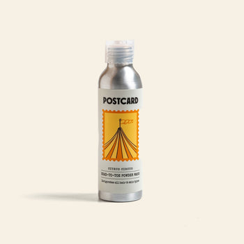 A silver bottle labeled Postcard Head-To-Toe Powder Wash, 80g - Citrus Circus, features a yellow circus tent in a stamp-like border and holds a citrus-infused powder-to-foam cleanser with vitamin C for refreshing hair and skin care.
