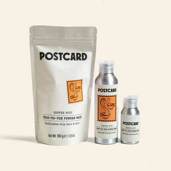 The Postcard Head-To-Toe Powder Wash, 80g - Coffee Spot features two silver bottles and a white pouch with orange duck stamp designs. This gentle powder-to-foam cleanser uses upcycled coffee grounds for eco-friendly skincare.