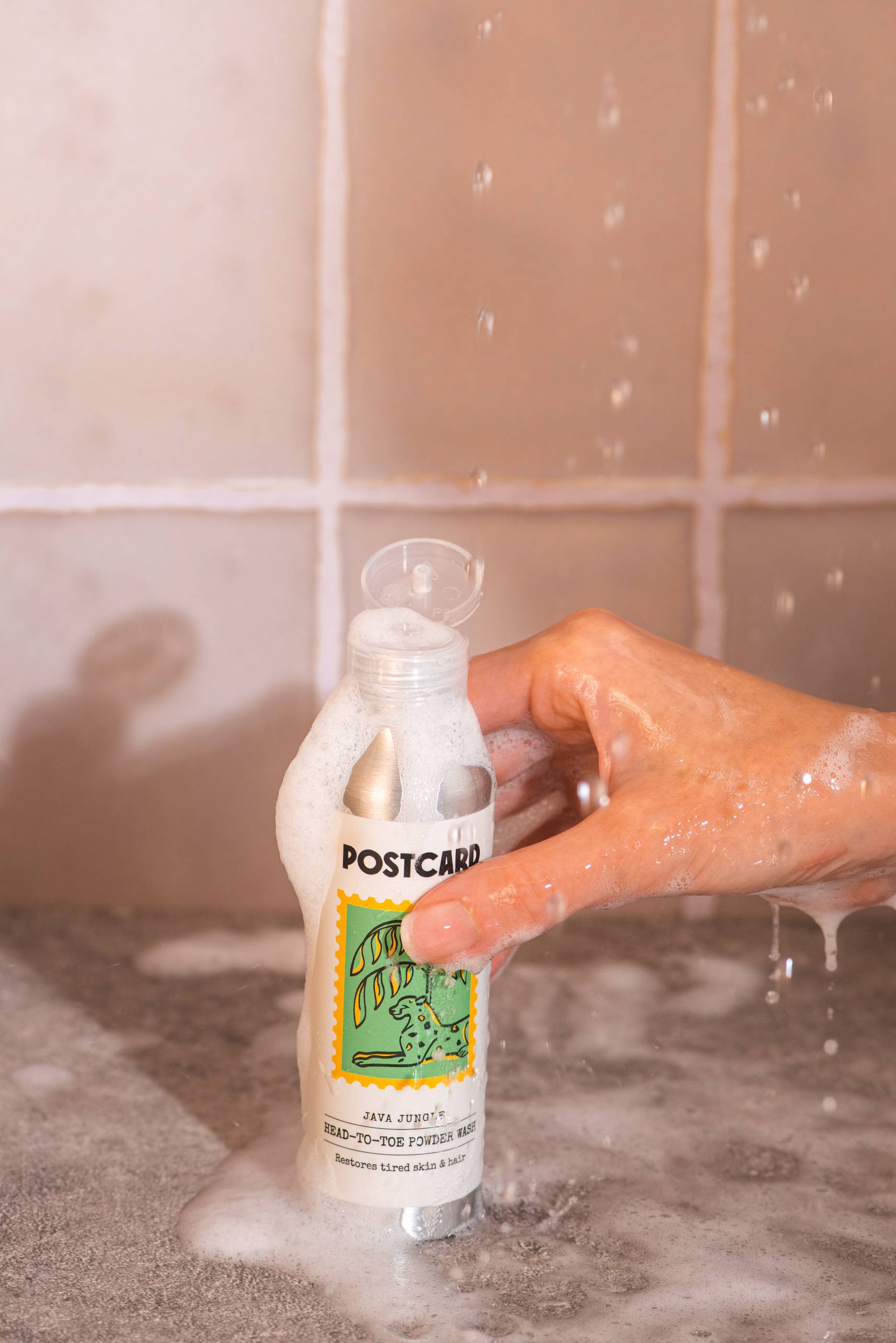 A hand with soap suds holds a bottle of Postcard Head-To-Toe Powder Wash, 80g - Java Jungle under running water, with tiles in the background.