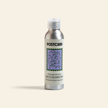 A silver 80g Postcard Head-To-Toe Powder Wash bottle, Woodland Retreat scent, with a purple floral design, stands upright on a plain background, evoking a luxurious spa experience.