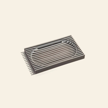 The Postcard Lattice Soap Dish by Postcard is a rectangular, dark gray soap dish made from recyclable resin with parallel slats and a central oval, perfect for a zero-waste lifestyle on any plain light surface.