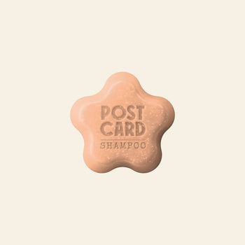 A beige, flower-shaped Postcard Shampoo Bar (60g - Citrus Circus) with POSTCARD SHAMPOO engraved on top, vitamin C-rich and sulfate-free, is set against a plain, light background. Brand: Postcard.
