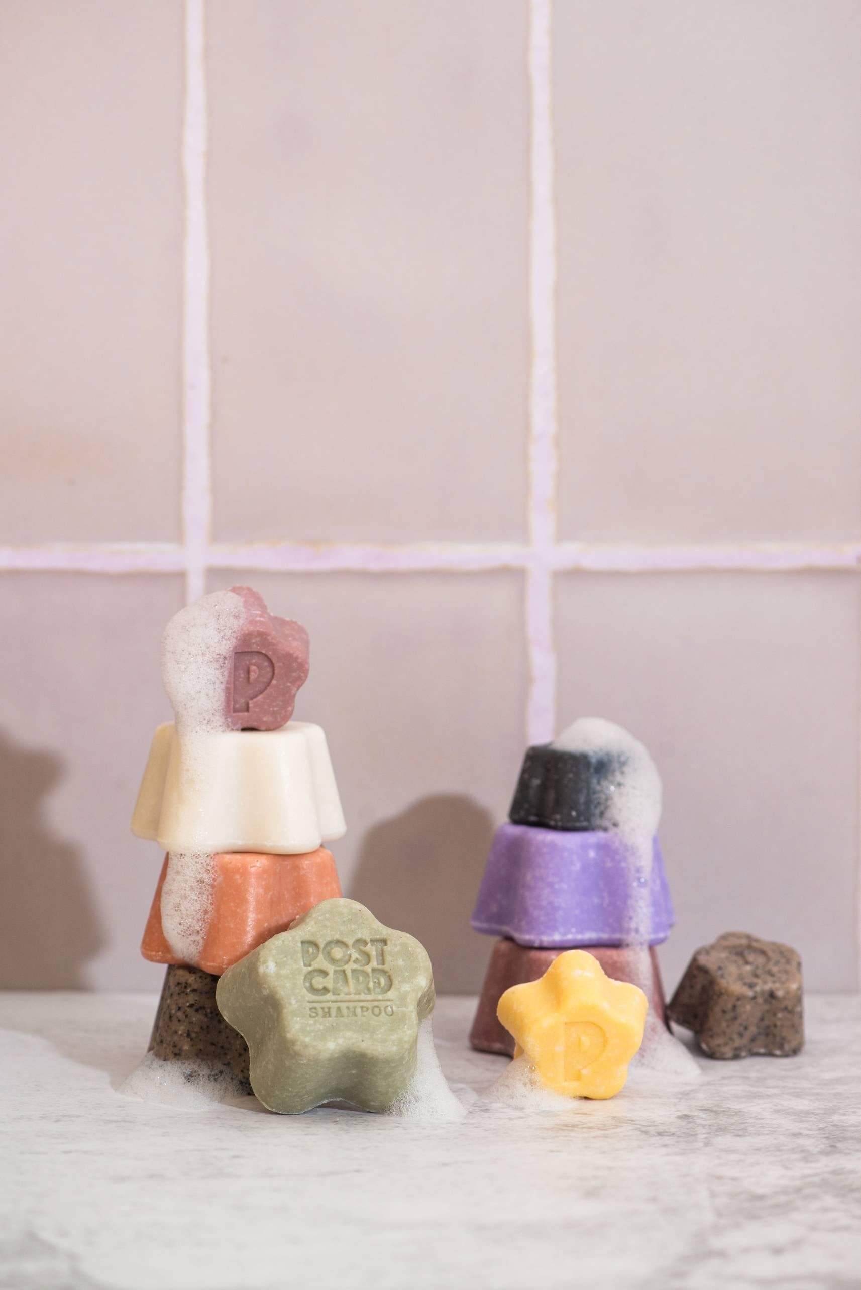 Colorful Postcard Shampoo Bars, like the 60g Citrus Circus, are sulfate-free and vitamin C-enriched. Featuring unique star and square shapes with playful bubbles, they’re displayed stacked on a wet surface before a tiled wall.