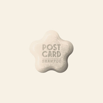 A flower-shaped Postcard Shampoo Bar, 60g – Clear Skies, embossed with POSTCARD SHAMPOO, rests on a light beige background. This sulfate-free bar from Postcard is ideal for sensitive scalps needing gentle, effective cleansing.