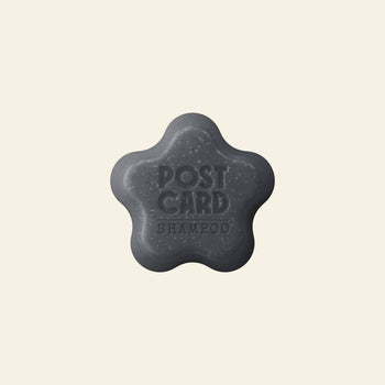 The Postcard Shampoo Bar, 60g - Icy Peaks, is a dark gray flower-shaped bar with activated charcoal for oily hair, embossed with POSTCARD SHAMPOO and displayed on a light beige background.