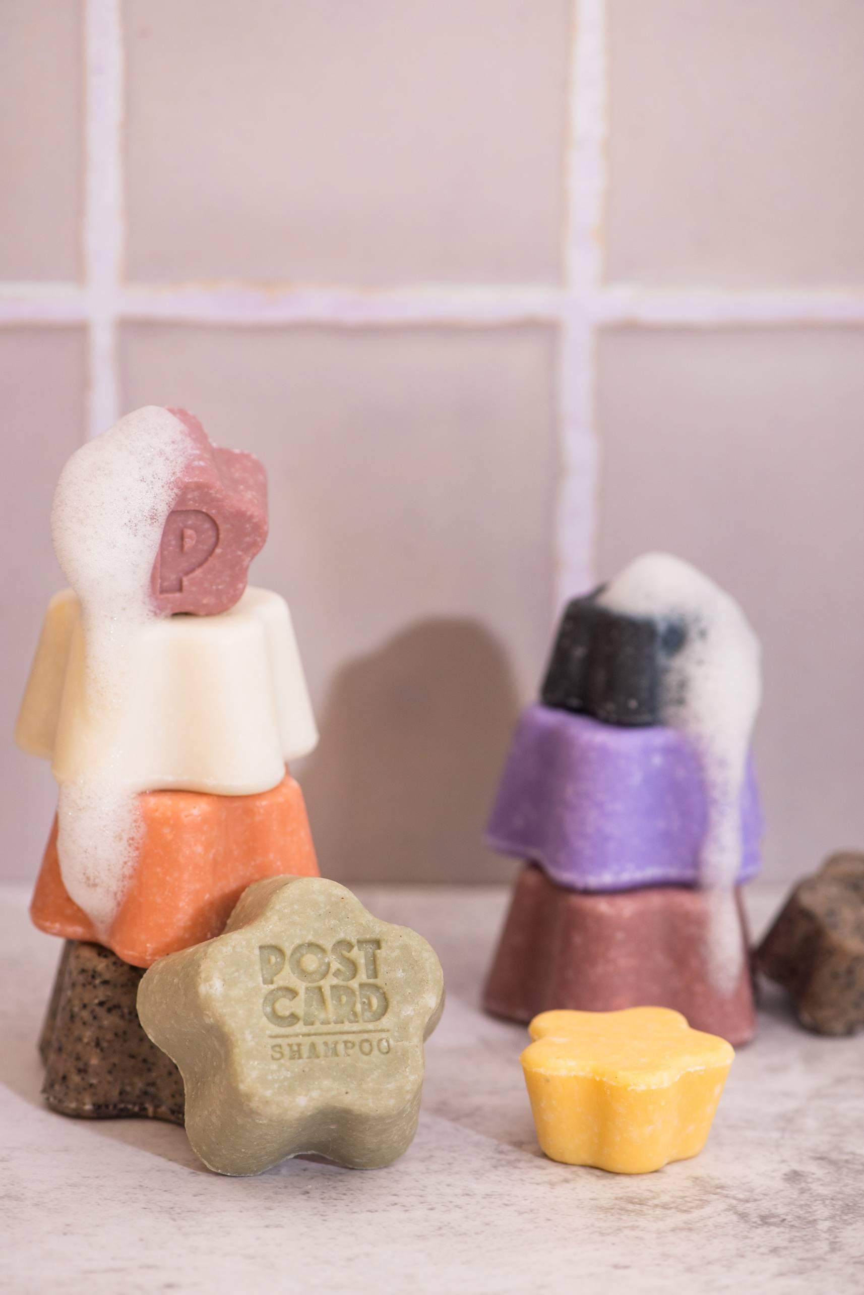 Colorful stacked soap bars, some bubbly, rest before a tiled wall. In front, a green Postcard Shampoo Bar—Java Jungle, 60g, sulfate-free—and a small yellow bar promise gentle cleansing and care.
