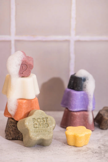 Colorful stacked soap bars, some bubbly, rest before a tiled wall. In front, a green Postcard Shampoo Bar—Java Jungle, 60g, sulfate-free—and a small yellow bar promise gentle cleansing and care.