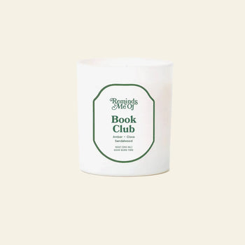 The Reminds Me Of Book Club Candle (10oz) by Reminds Me Of is a white candle in a glass jar with warm sandalwood, clove, and amber notes, shown against a plain light background.