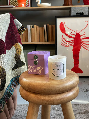 A round wooden stool holds a purple Reminds Me Of Book Club Candle, 10oz, scented with amber and sandalwood. Behind it are wooden shelves with books, a colorful blanket, and framed art featuring a red lobster.
