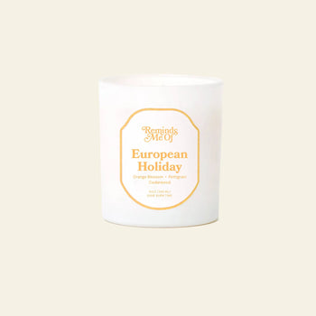 The Reminds Me Of European Holiday Candle (10oz) by Reminds Me Of is a white candle in a glass jar with notes of orange blossom, bergamot, pomegranate, and cedarwood, shown against a plain beige background.