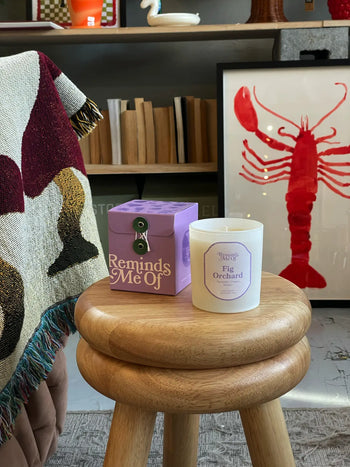 A wooden stool displays a purple Reminds Me Of Fig Orchard Candle, 10oz by Reminds Me Of. A colorful blanket hangs nearby, with shelves and red lobster artwork visible in the background.