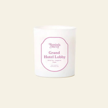 The Reminds Me Of Grand Hotel Lobby Candle, 10oz features uplifting scents of White Tea, Bergamot, and Ginger. This white candle with a pink label is set against a plain light beige background. Brand: Reminds Me Of.