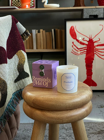 A wooden stool holds a Reminds Me Of Poolside Candle, 10oz, in purple and coconut milk scent, next to a patterned blanket and framed red lobster artwork on a bookshelf.