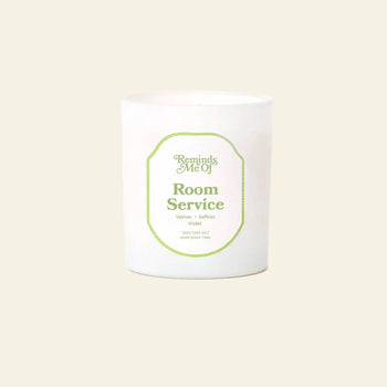 A 10oz Reminds Me Of Room Service Candle by Reminds Me Of, in a simple glass jar with soothing vetiver and violet notes, is displayed against a plain beige background.
