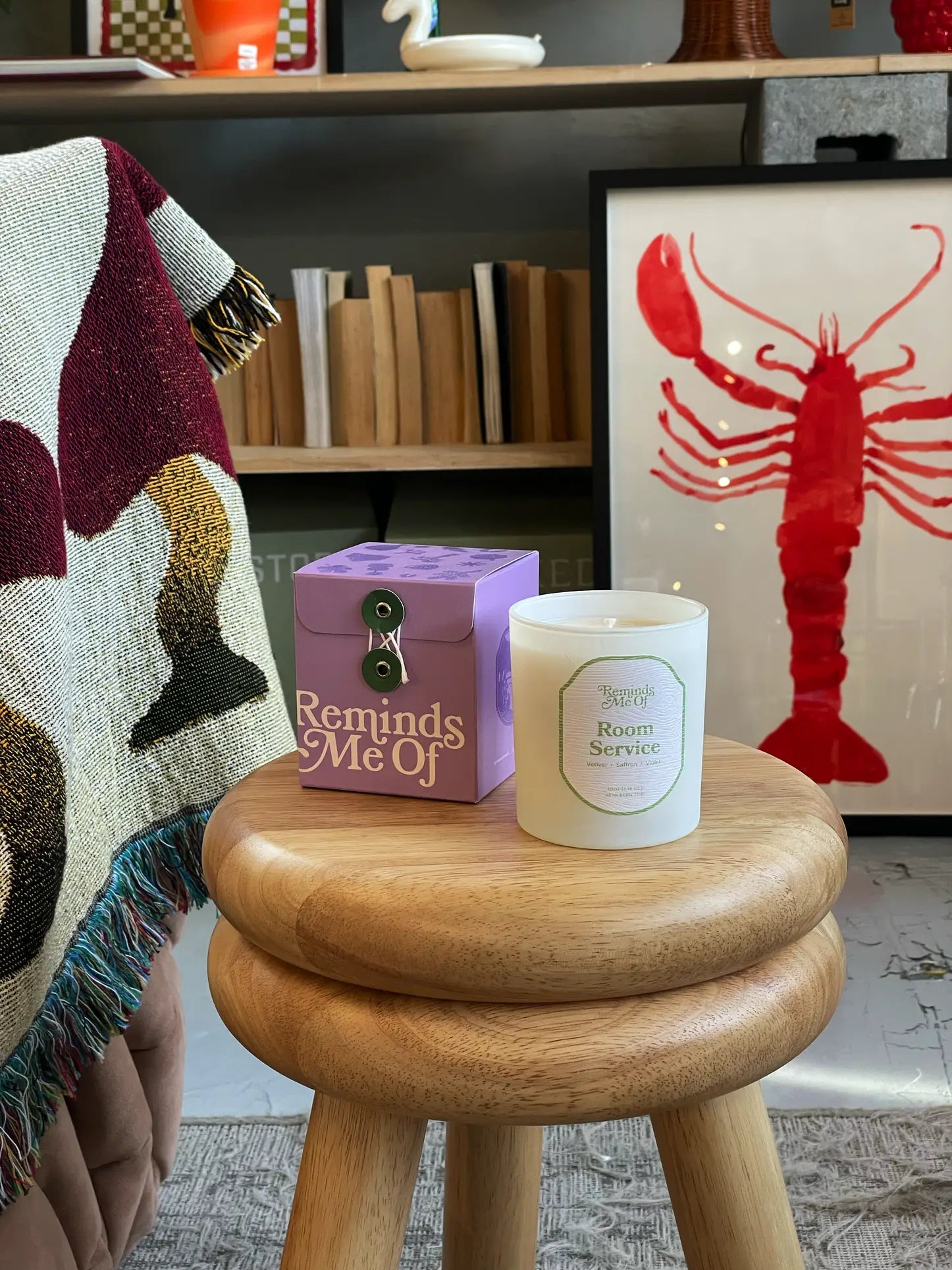 A violet Reminds Me Of Room Service Candle (10oz) sits on a wooden stool beside its box. Behind them are a fringed blanket, bookshelf, and a large framed red lobster print.