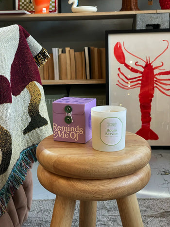 A violet Reminds Me Of Room Service Candle (10oz) sits on a wooden stool beside its box. Behind them are a fringed blanket, bookshelf, and a large framed red lobster print.