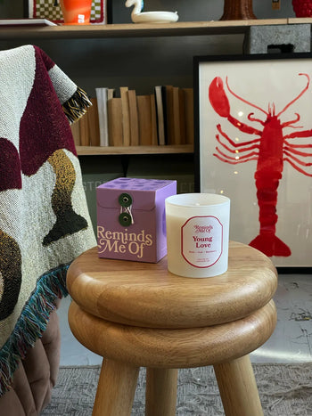 A 10oz Reminds Me Of Young Love Candle rests on a wooden stool in its purple box, surrounded by red lobster art, books, and a patterned blanket draped over furniture.