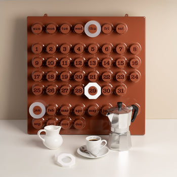 A Ring-A-Date Limited Edition Wall Calendar - Cafè Mousse is mounted on a beige wall. In front, there's a stovetop espresso maker, a coffee cup with saucer, and a small white milk jug.