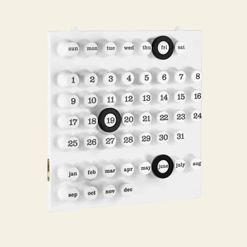 The Ring A Date Wall Calendar by Ring-A-Date is a modern, white Italian calendar with raised ABS plastic buttons for days, dates, and months. Black rings highlight Friday the 19th and June on a minimalistic layout with a plain light background.