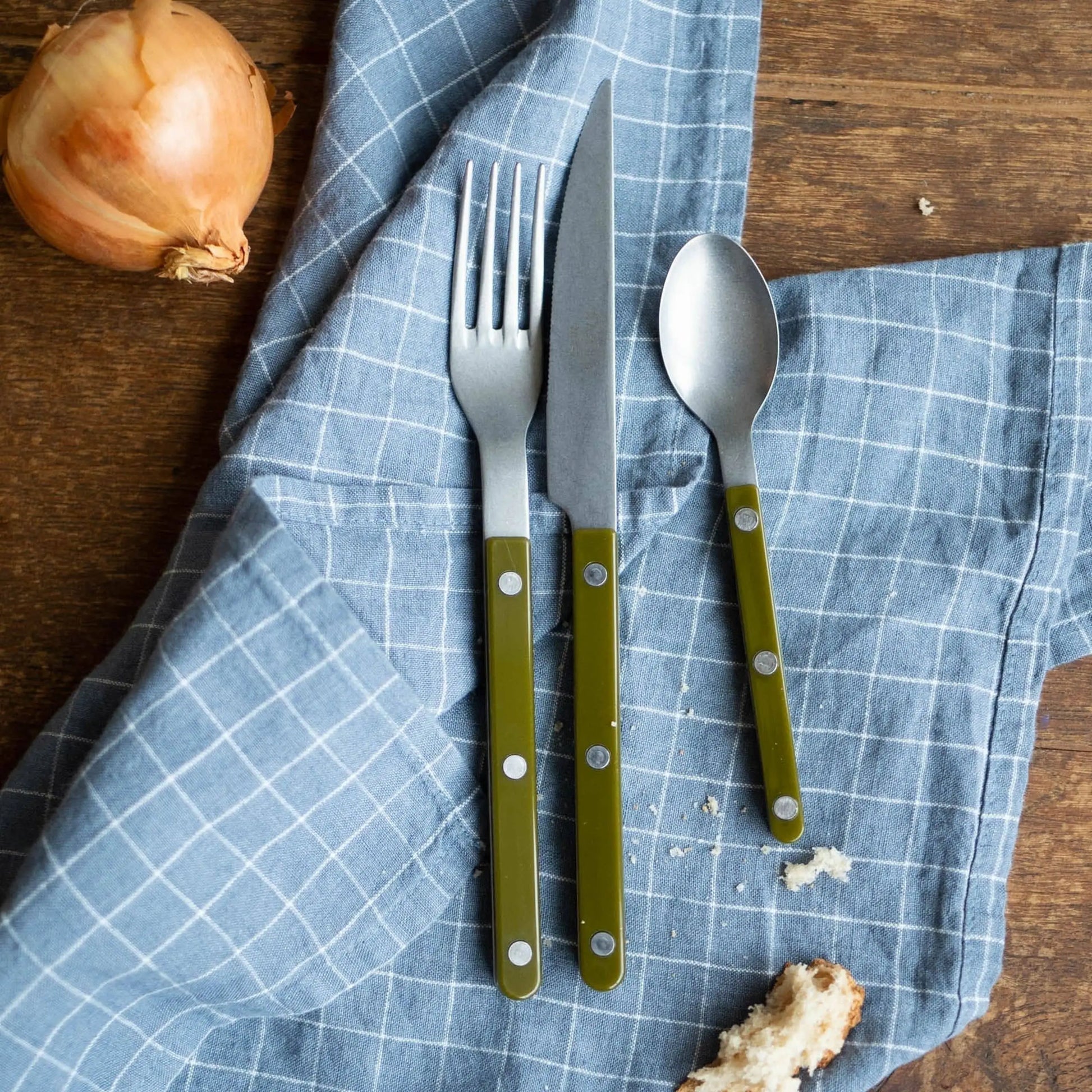 A fork, spoon, and Sabre Bistrot Vintage Dinner Knife with green handles rest on a blue checkered cloth on a wooden table beside an onion and a piece of bread.