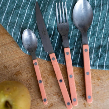A Sabre Bistrot Vintage Dinner Knife with a pink handle is arranged with a small spoon, a fork, and a large spoon on a wooden surface next to a green striped cloth and a pear.