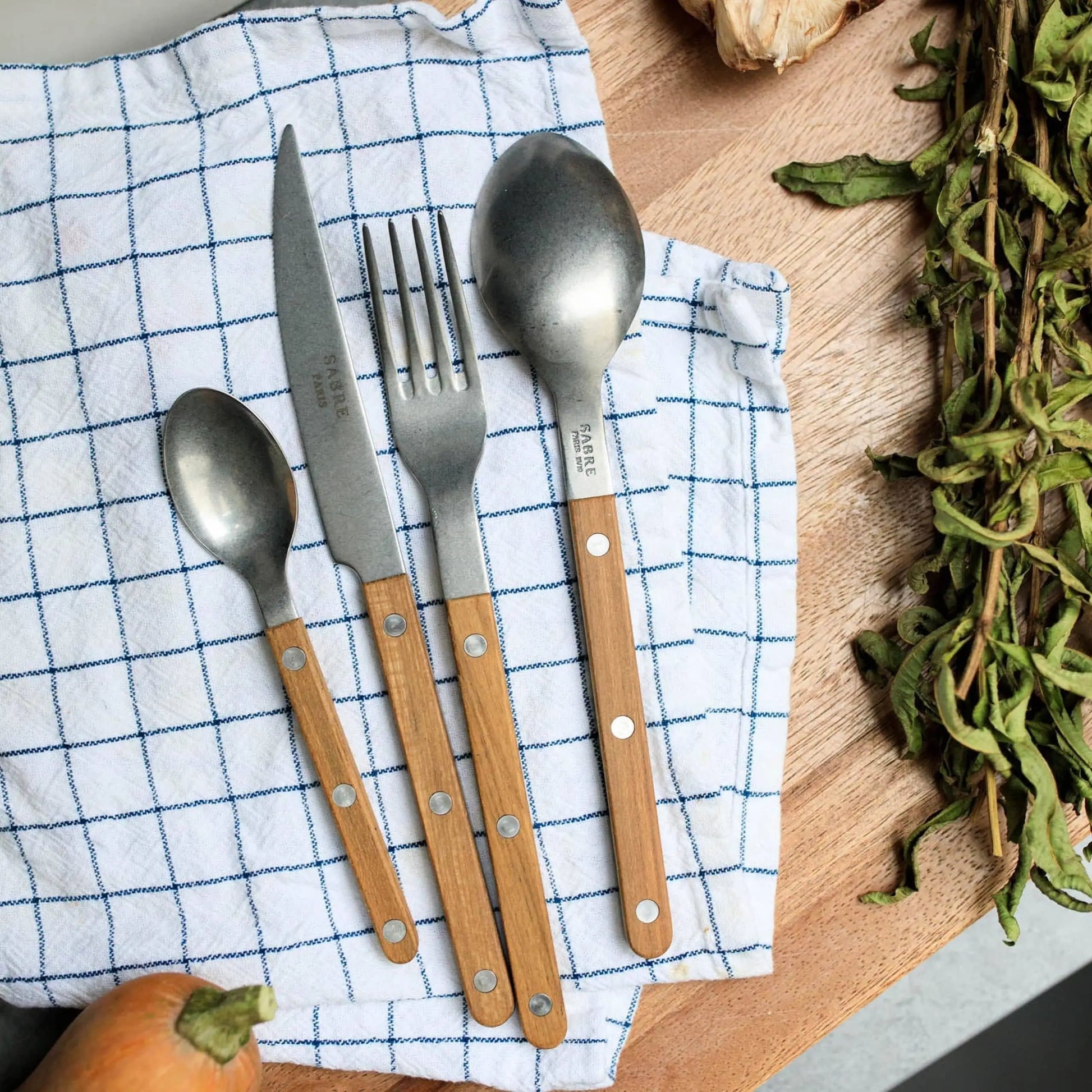 A set of four utensils with wooden handles—small spoon, fork, large spoon, and a Sabre Bistrot Vintage Dinner Knife—rests on a blue-and-white checkered cloth beside leafy greens on a wooden surface.