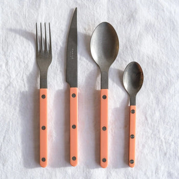 The Sabre Bistrot Vintage Soup Spoon by Sabre, with a matte stainless steel finish and a light pink handle, is arranged alongside three other matching cutlery pieces on a white textured fabric background.