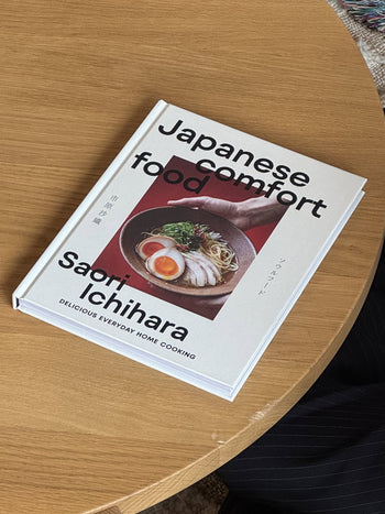 Saori Ichihara Japanese Comfort Food by Saori Ichihara rests on a wooden table, its cover featuring ramen with meat, eggs, and green onions—inviting you to discover authentic Japanese recipes.