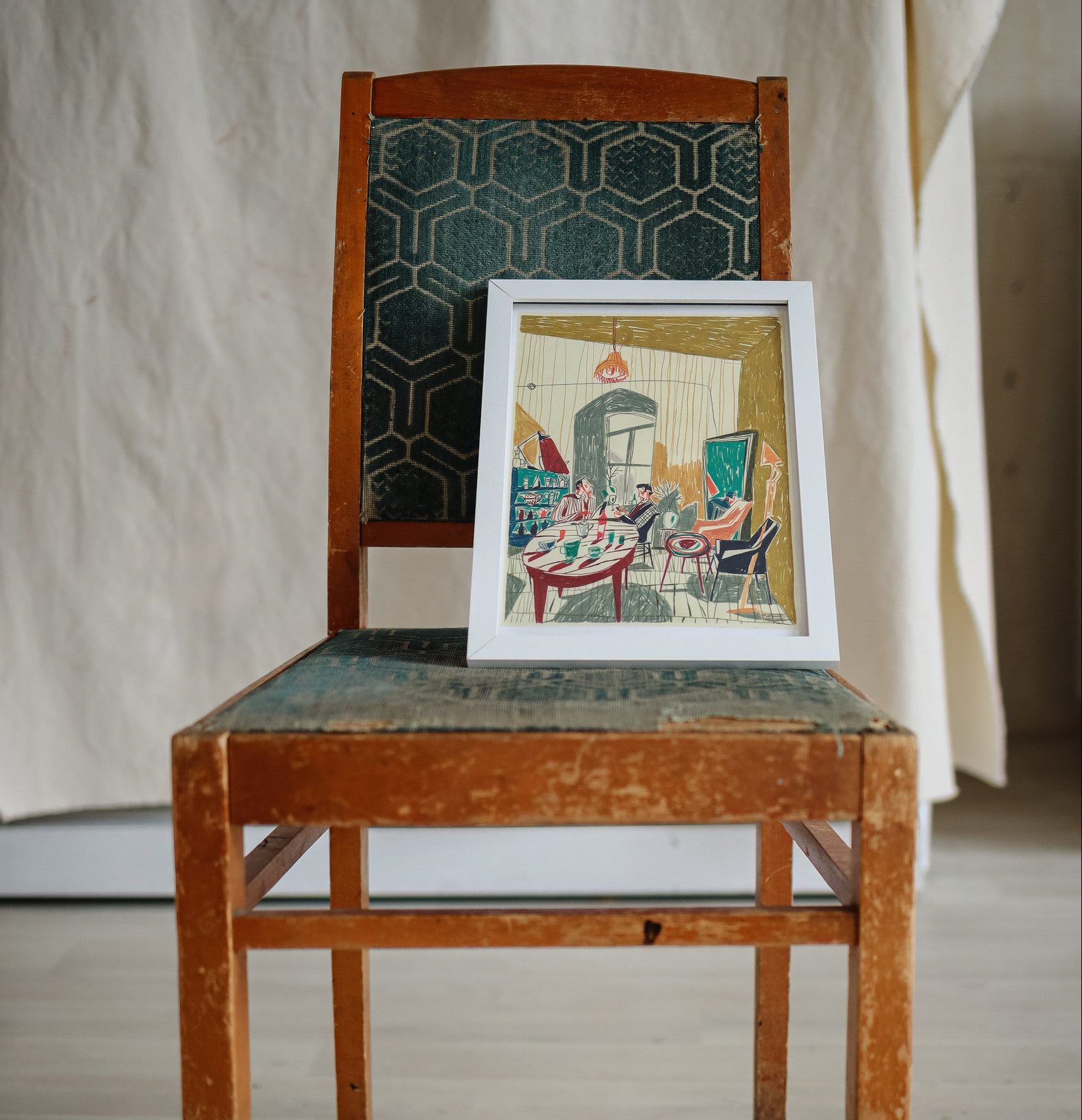 The Evening at Mirzoyan Librarys Caffe print by Sarah Mari Shaboyan rests on a vintage wooden chair with a green cushion, evoking the charm of tea at Mirzoyan Library against a neutral fabric backdrop.