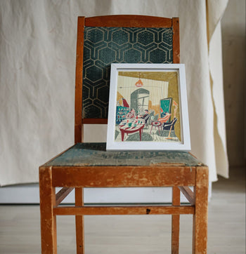 The Evening at Mirzoyan Librarys Caffe print by Sarah Mari Shaboyan rests on a vintage wooden chair with a green cushion, evoking the charm of tea at Mirzoyan Library against a neutral fabric backdrop.