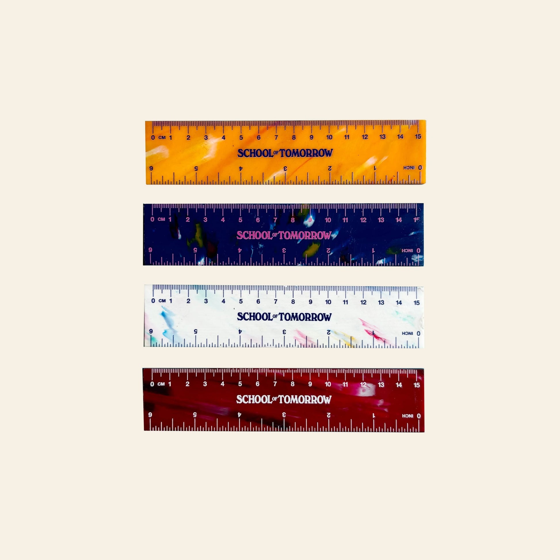 Four 15cm School of Tomorrow rulers, made from recycled bottle caps, display a unique design in orange, navy blue, white, and red—each labeled SCHOOL TOMORROW. They are arranged horizontally on a light beige background.