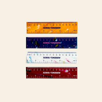 Four 15cm School of Tomorrow rulers, made from recycled bottle caps, display a unique design in orange, navy blue, white, and red—each labeled SCHOOL TOMORROW. They are arranged horizontally on a light beige background.