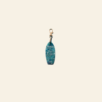 The School of Tomorrow Keychain - Blue by School of Tomorrow is a small surfboard-shaped keychain, handmade from repurposed plastic, with BEACH, an ocean wave design, and a gold clasp displayed on a plain beige background.