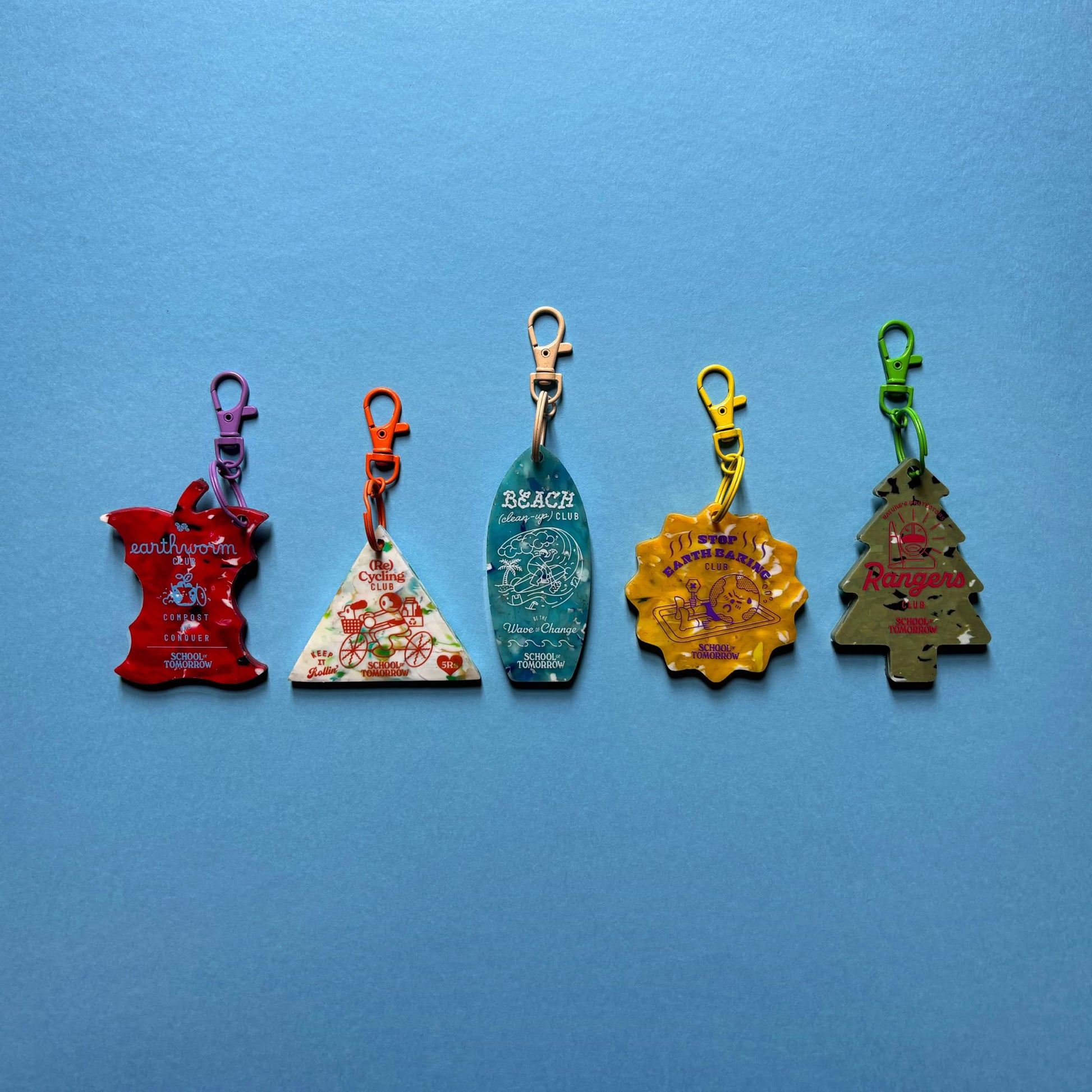 The School of Tomorrow Keychain - Green by School of Tomorrow, made from recycled HDPE plastic, is displayed among five colorful, uniquely shaped handmade keychains on a blue background.