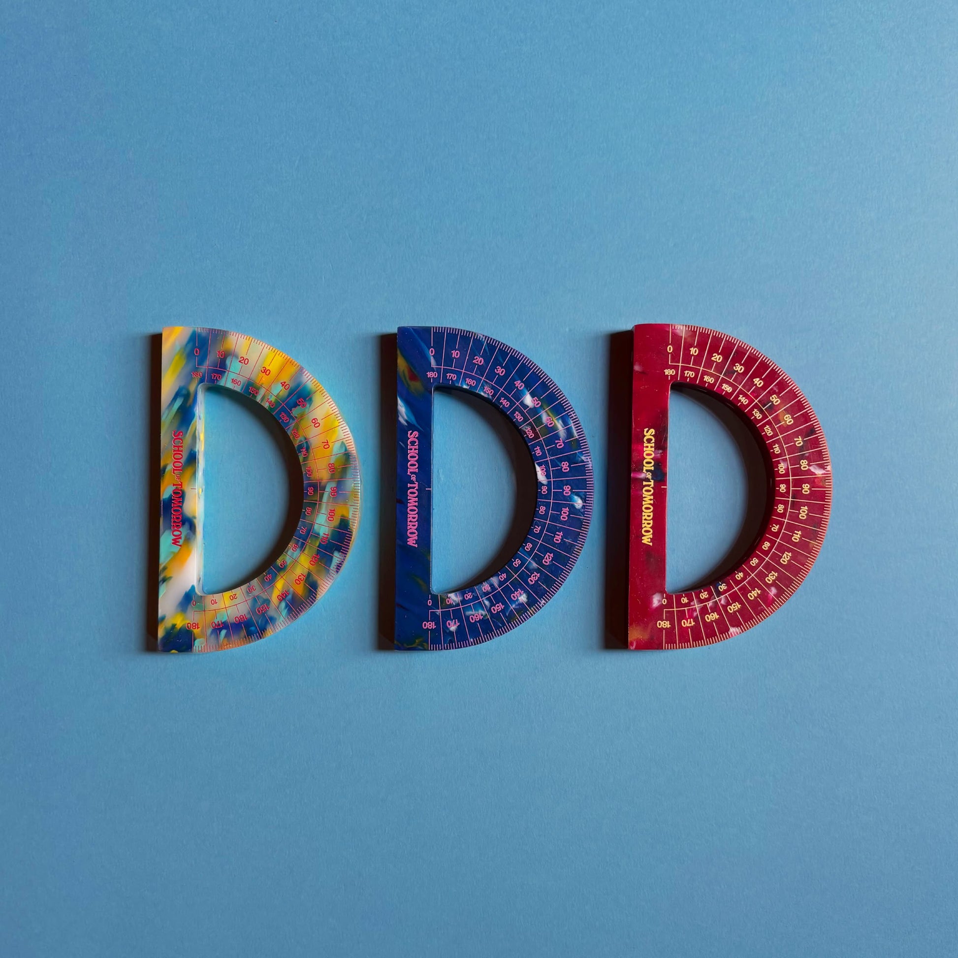 Three School of Tomorrow Math Instruments Protractors—multicolored and eco-friendly, made from recycled HDPE—are arranged side by side on a blue background to form the letters DDD in yellow, blue, and red.