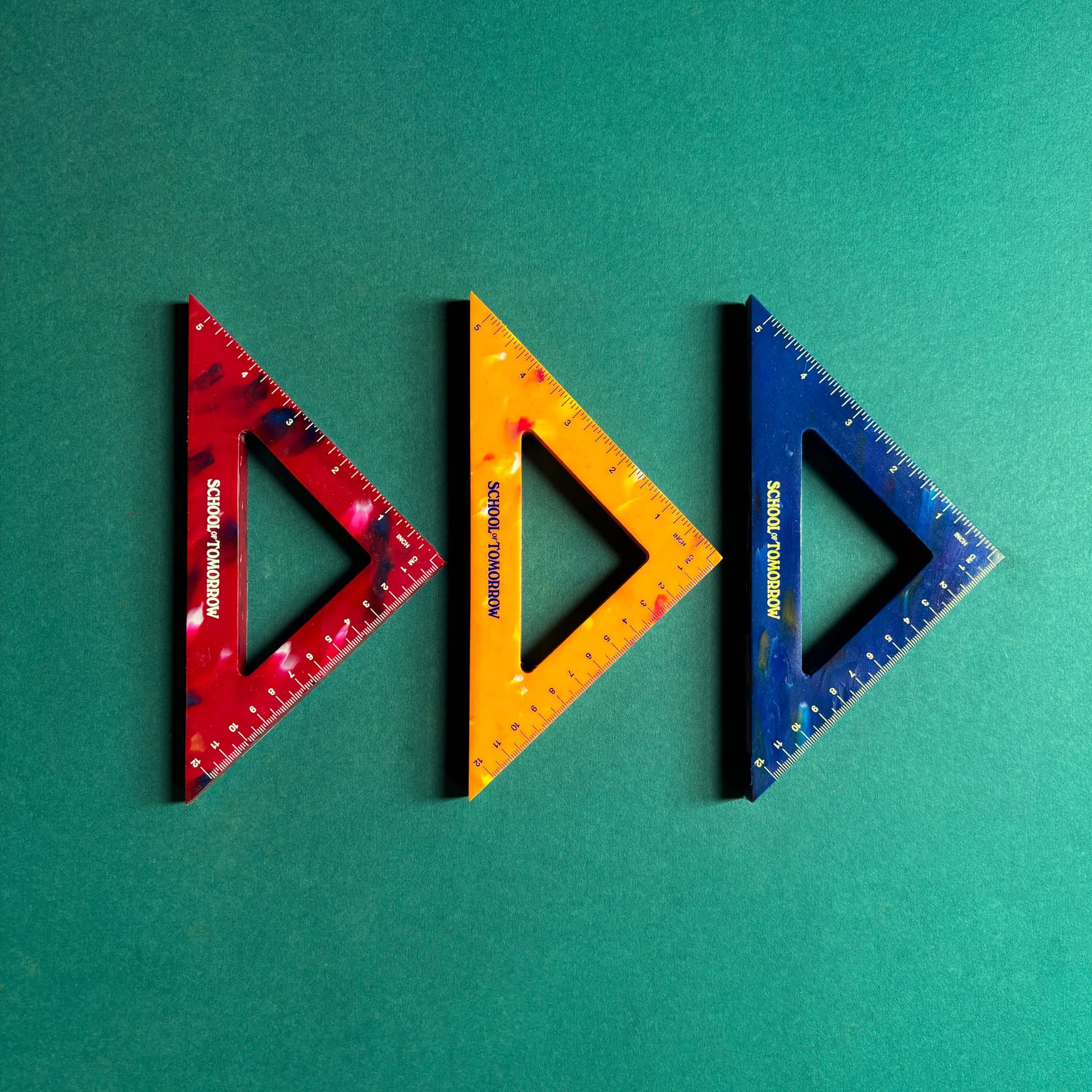 Three triangular rulers in red, yellow, and blue from School of Tomorrow are side by side on a green background. Made from recycled bottle caps, these eco-friendly triangles have measurement markings and the School of Tomorrow logo.