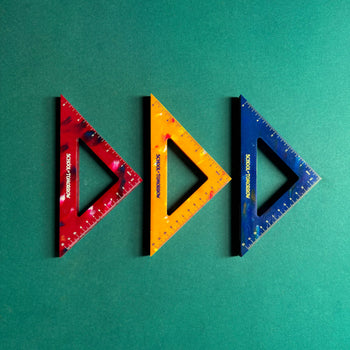 Three triangular rulers in red, yellow, and blue from School of Tomorrow are side by side on a green background. Made from recycled bottle caps, these eco-friendly triangles have measurement markings and the School of Tomorrow logo.
