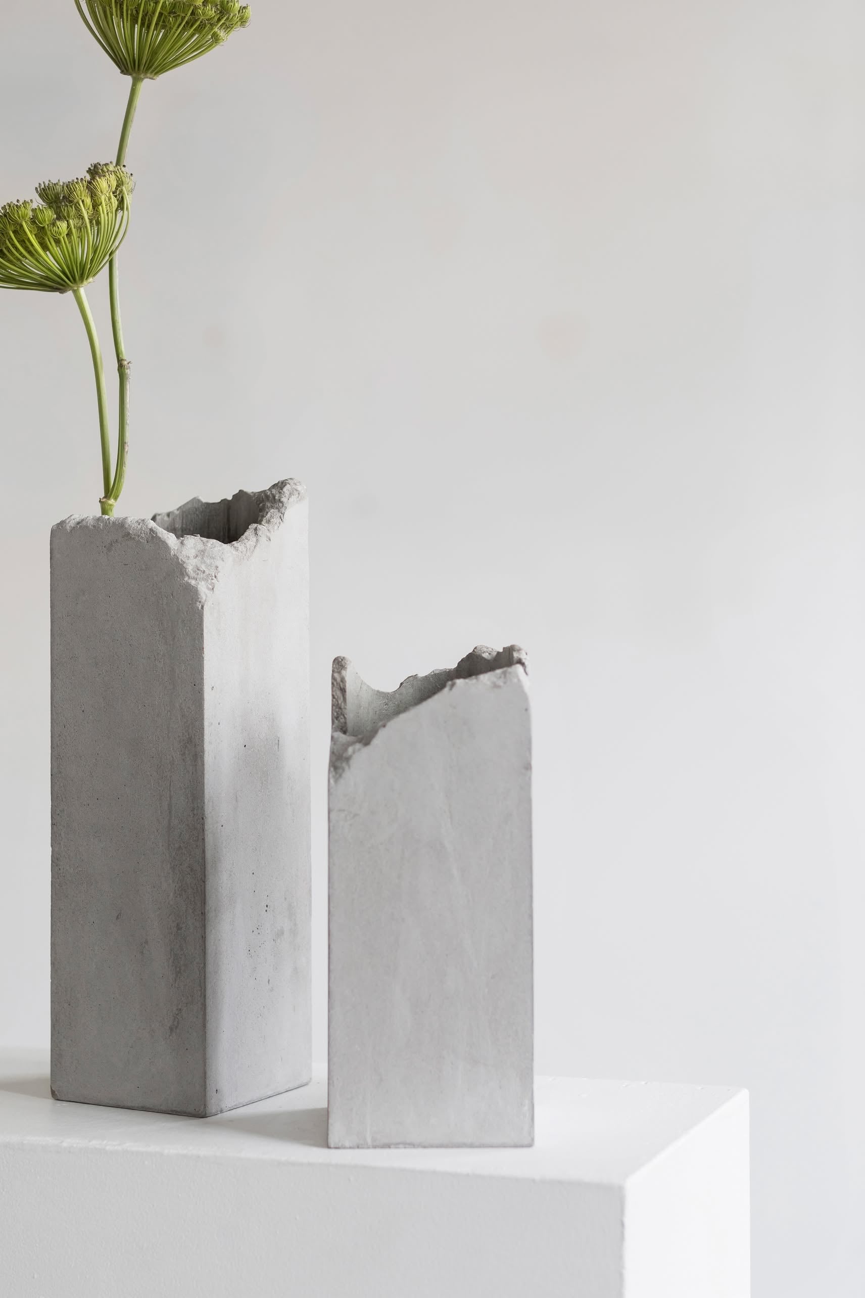 Two Serax Broquaine Concrete Vases S - Grey by Serax, with rough, uneven tops, rest on a white surface. One holds a single green flower stem, highlighting modern design against a plain light background.