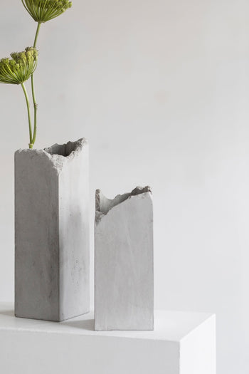 Two Serax Broquaine Concrete Vases S - Grey by Serax, with rough, uneven tops, rest on a white surface. One holds a single green flower stem, highlighting modern design against a plain light background.