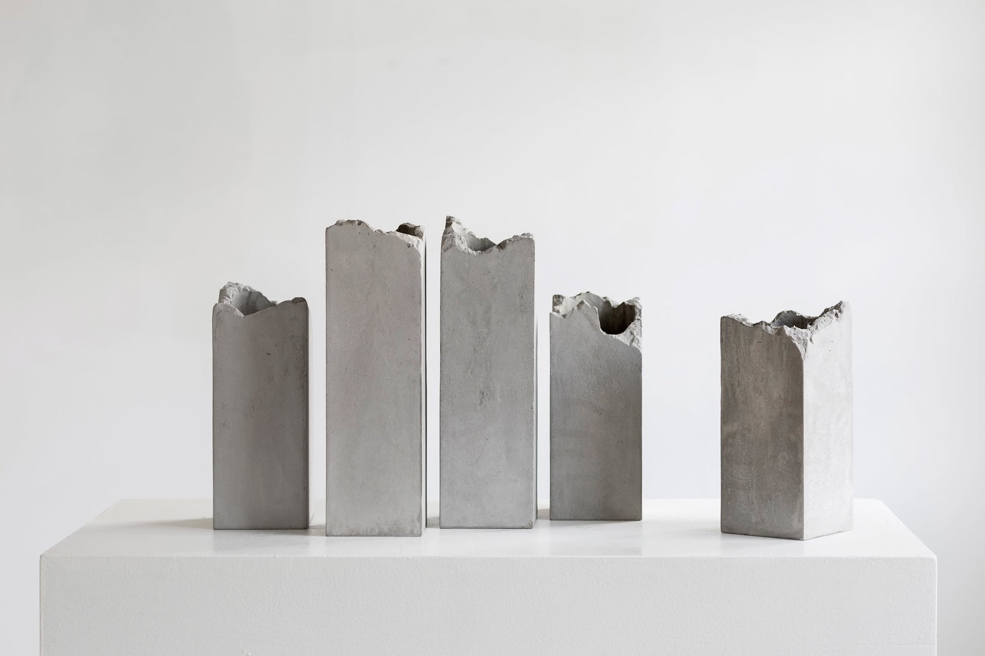Five upright, rectangular Serax Broquaine Concrete Vase S - Grey by Serax stand on a white pedestal against a plain background. Their jagged tops suggest modern artistry and highlight the unique character of the collection.