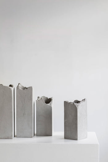Four tall, rectangular Serax Broquaine Concrete Vase S - Grey by Serax are displayed on a white pedestal against a light gray background, their jagged tops evoking minimalist, industrial sculpture reminiscent of modern artistry.