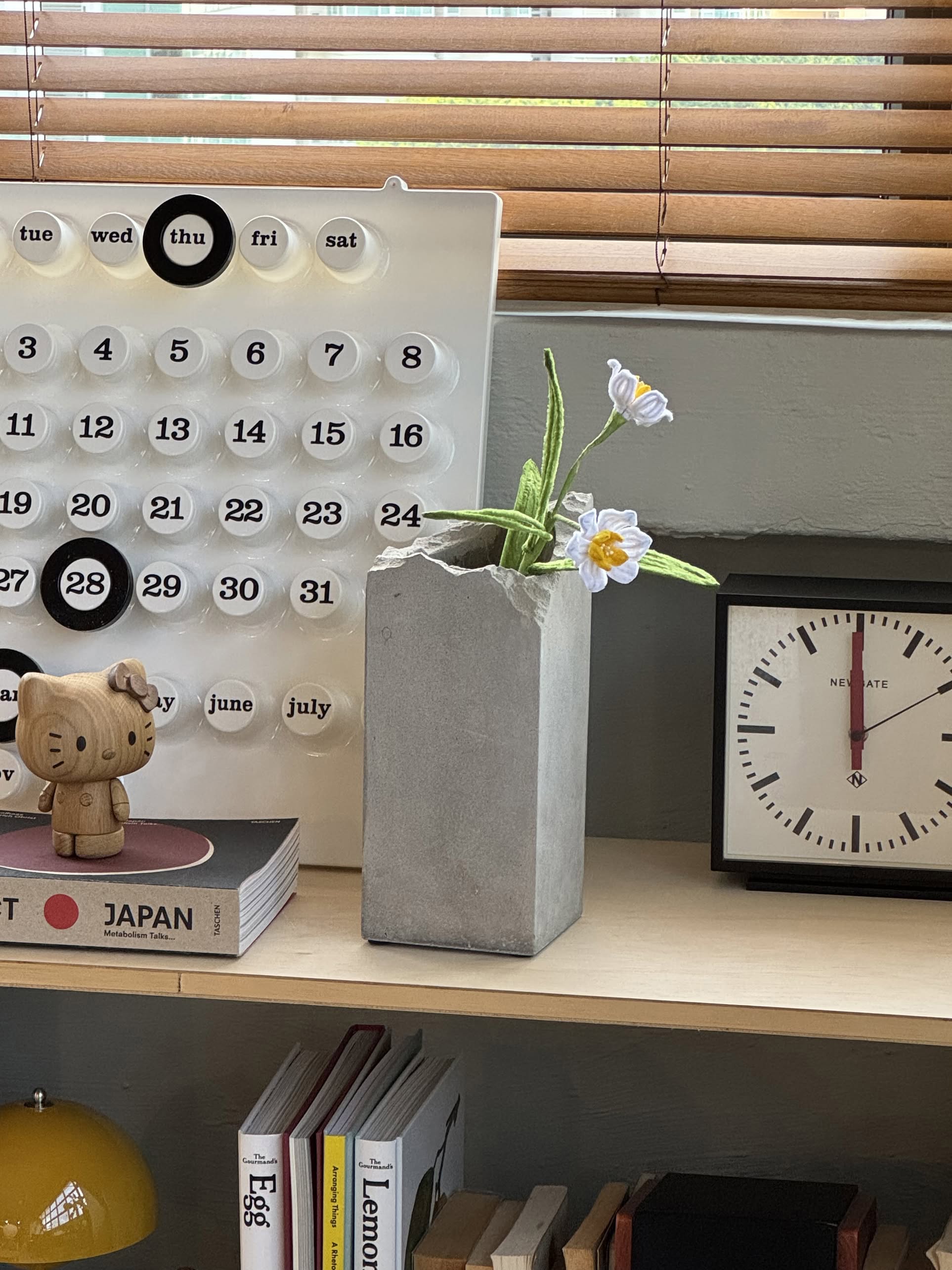 A desk with a modern calendar, a small clock, the Serax Broquaine Concrete Vase S in grey by Serax holding white and yellow flowers, a wooden figurine, and books stacked neatly on a shelf below.