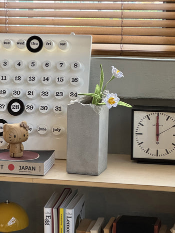 A desk with a modern calendar, a small clock, the Serax Broquaine Concrete Vase S in grey by Serax holding white and yellow flowers, a wooden figurine, and books stacked neatly on a shelf below.