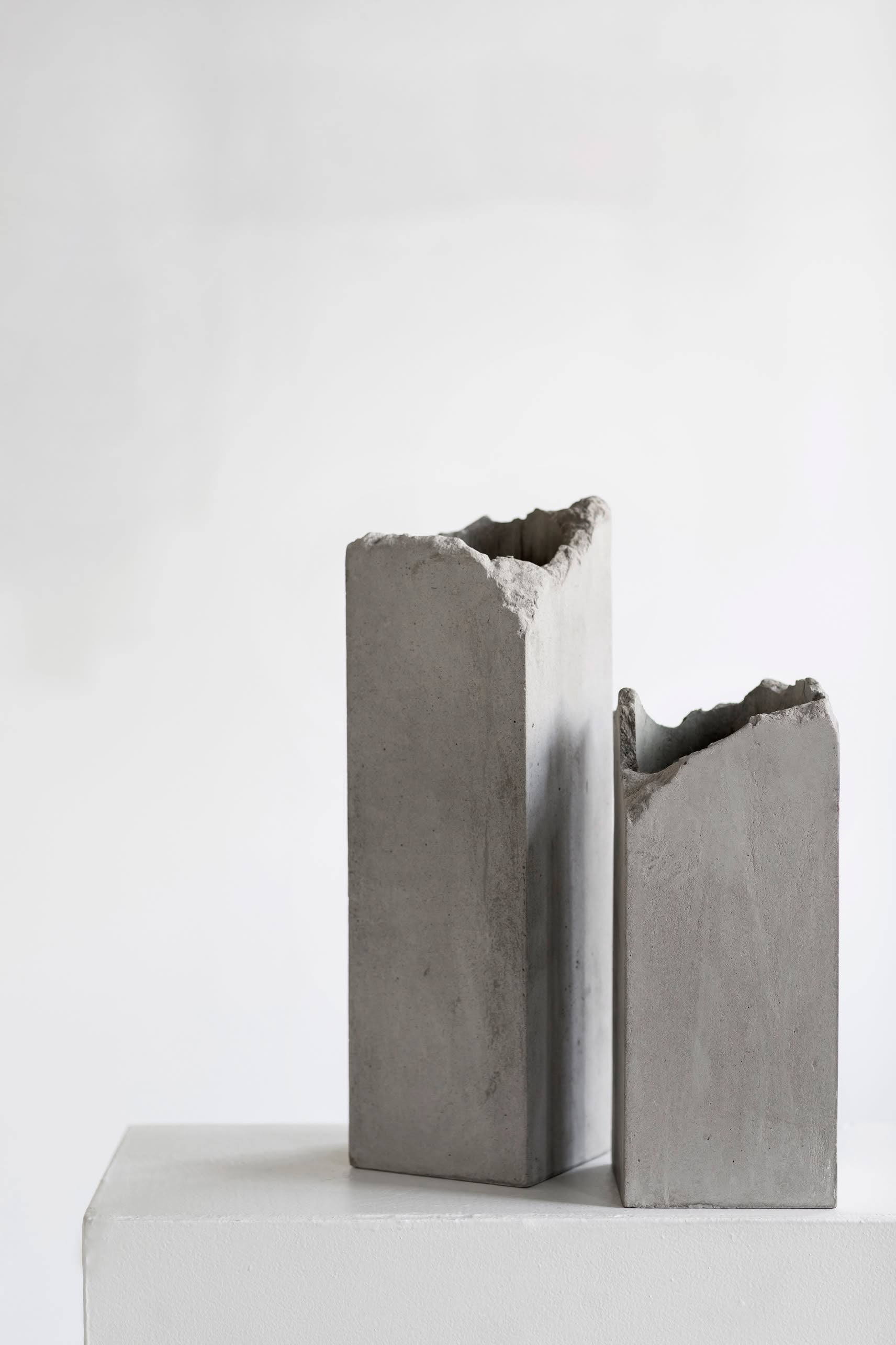 Two Serax Broquaine Concrete Vases S - Grey by Serax, with minimalist rectangular forms and jagged tops, are displayed side by side on a white pedestal against a light gray background.